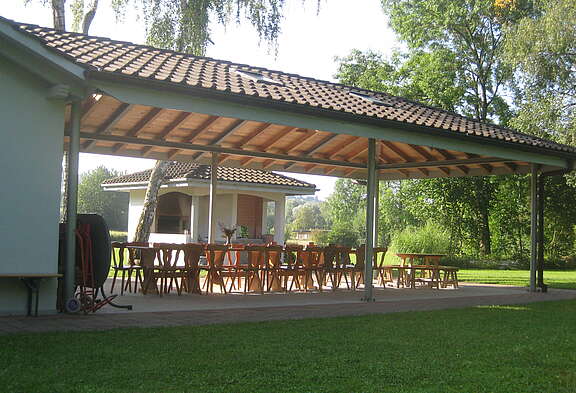 Überdachter Grill- und Essplatz des Seehauses Mörigen mit Holztischen und -stühlen inmitten einer grünen, ruhigen Gartenanlage.